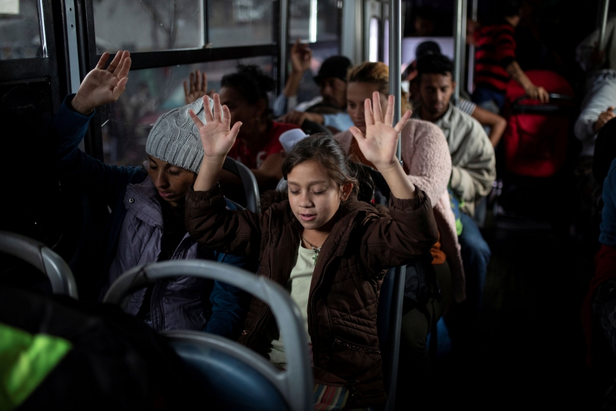 Migrants pray before moving by buses to a new shelter. Migrants are shown with their hands raised in the air praying on a bus.