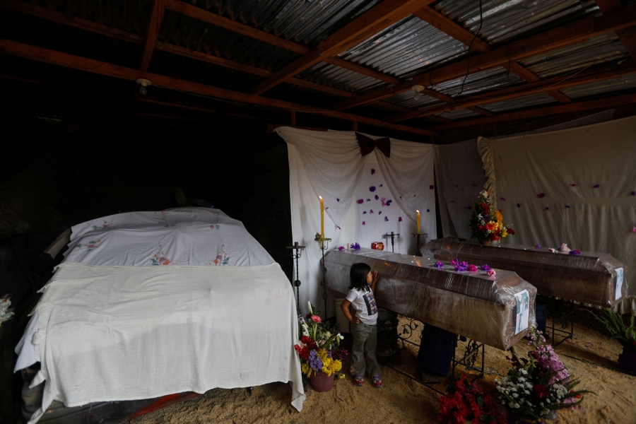 A girl stands next to the coffins containing the bodies of Blanca and another victim of the violence during a wake inside a garage, in Tegucigalpa, Honduras. A young girl is shown standing next to two wooden coffins wrapped in plastic wrap.