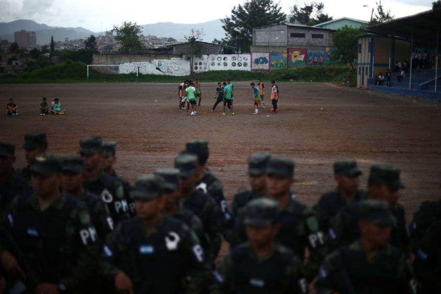 People play soccer as soldiers line up in a neighborhood occupied by gangs known as El Hoyo in Tegucigalpa, Honduras. A group of people play soccer in the distance as soldiers are shown in the near ground lined up.