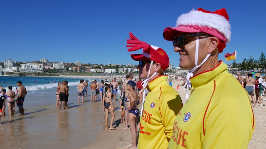 Volunteer lifeguards wearing yellow jackets and red and white caps on Christmas Two men wearing Santa Claus's hats stand on a sunny beach surrounded by people in bathing suits in Sidney Australia.