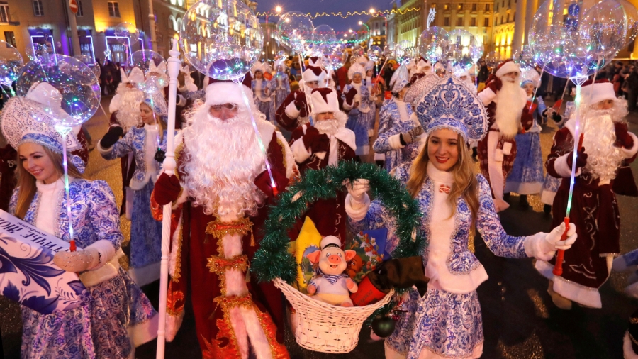 Father Frost and Snow Maiden in Belarus. Men dressed in red and women dressed in blue and white Christmas attire walk through streets holding Christmas lights.
