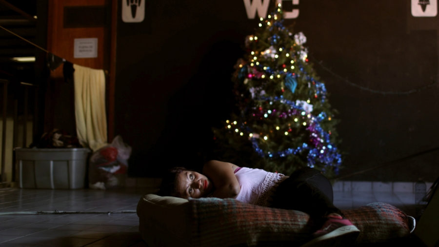 Migrant in Tijuana on Christmas. A woman lays on a mattress on the floor in front of a Christmas tree in a dark room in a shelter in Tijuana, Mexico.