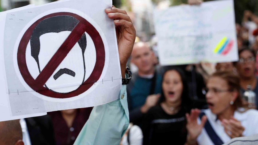 A hand holds up a poster with a simple icon of a man with a mustache in a red "no" circle