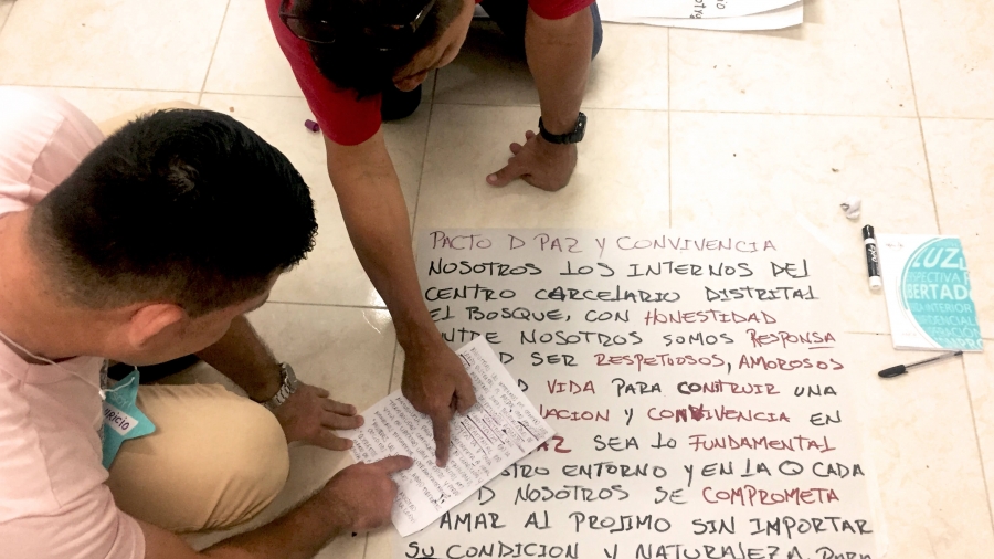 Two men sit on the floor and lean over a piece of paper with writing on it.