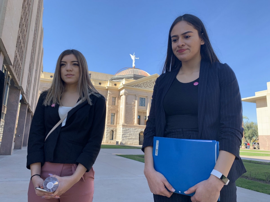 Two young people stand in front of the camera with a Capitol building behind them