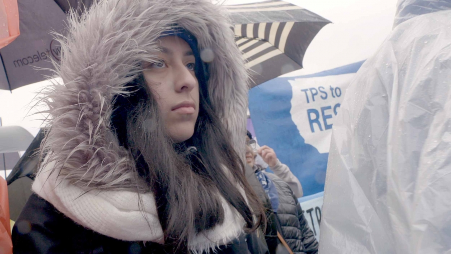 A close-up photo of a young woman at a TPS protest. A close-up photo of a young woman wearing a furry hood at a TPS protest with blue signs in the background.