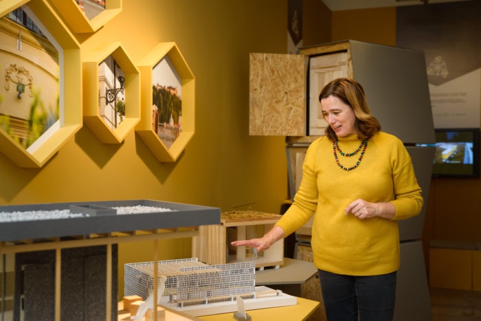 A woman stands over a small model of a beehive. Behind her is a yellow wall with honey-comb shaped pictures.