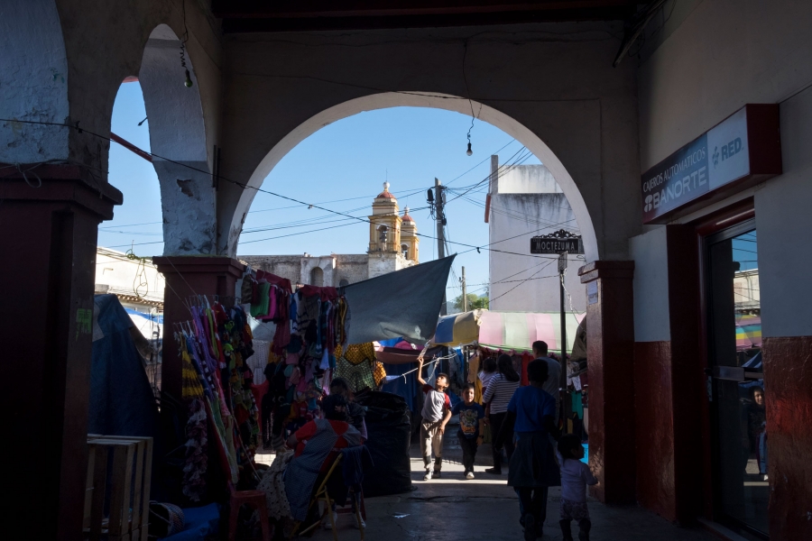 Locals mingle at Tlaxiaco’s market, with the Santa Maria de la Asunción church in the distance. People are shown walking around at Tlaxiaco’s market, with the steeples of the Santa Maria de la Asunción church in the distance.