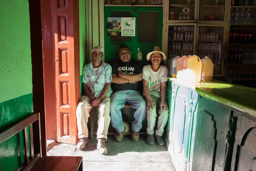 Enrique Reyes Rodríguez, 49, (center) went to Los Angeles in 1989 in search of work. He returned nine years later to visit his parents, and met his now-wife. He frequently listens to "La voz de la Mixteca." Three men are shown sitting on a bench in a store with bottles on the wall.