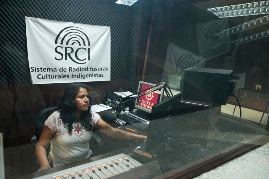 Eva Hernández, lead voice of “La Hora Mixteca” at La Voz de la Mixteca. Eva Hernández is shown sitting behind clear class at a radio DJ board with a microphone.