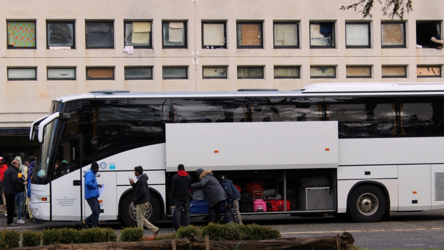 People load a white bus with luggage in front of a cement building.