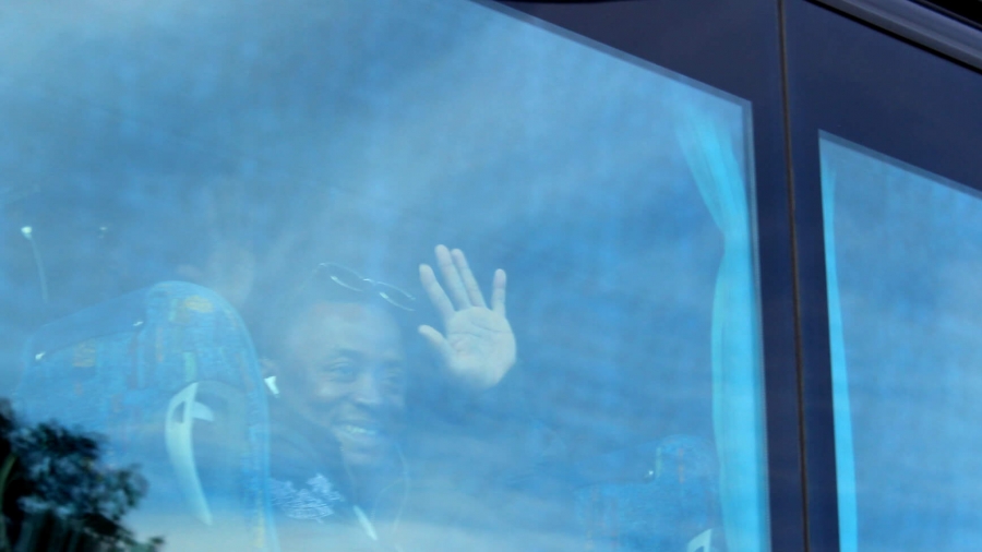 A young African man waves goodbye from the window on a bus.