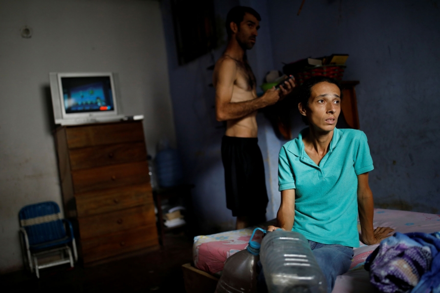 Guzman sits on her mother-in-law's bed as her husband, Jorge Perez, stands behind her. A woman is shown sitting a bed wearing a blue shirt as her husband stands behind her.