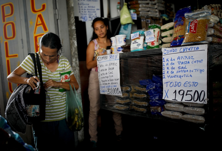 Guzman goes shopping for groceries. A woman is shown at a market with signs in Spanish for various groceries in front of her.