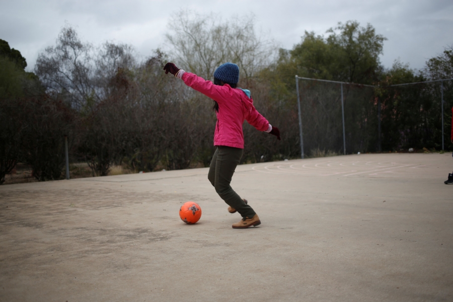 A young migrant plays with a ball at the monastery. A young girl wearing a pink jacket and wearing a winter hat plays with a ball.
