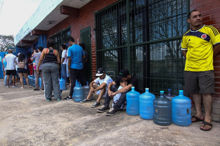People wait in line to try to buy potable water during a blackout in Puerto Ordaz, Venezuela. People a showning standing in line next to a brick building waiting with empty jugs.