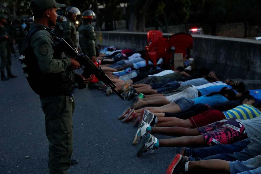 A security force member stands next to detainees on a street after looting during an ongoing blackout in Caracas, Venezuela. A security force member stands holding a rifle next to detainees laying on the street, face down.