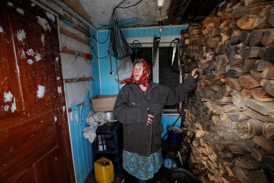 Taranenko looks at bullet holes in door caused by shelling and points at self-made protecting wall made of firewood in her house. A woman is shown with her hand on a wall of firewood looking to her right at bullet holes in door.