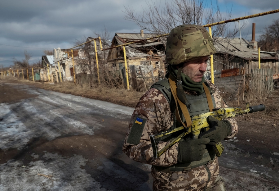 Oleg walks at the front line. A man in military fatiques walks the empty street carrying a rifle across his chest.