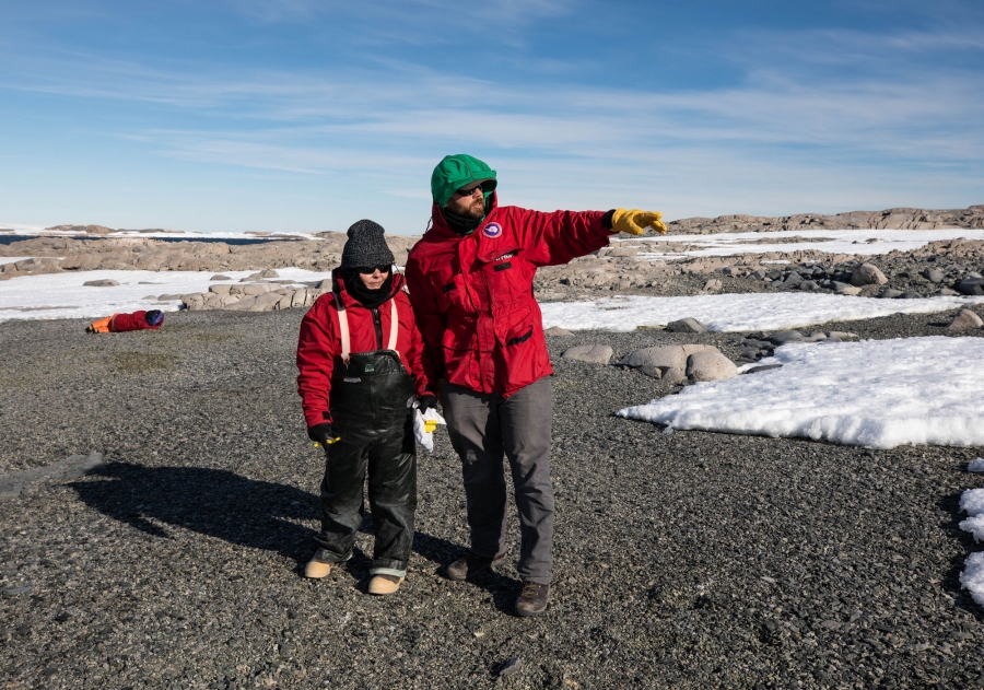 Victoria Fitzgerald and Scott Braddock are shown on one of the Schaefer Islands wearing heavy red jackets with Scott pointing off to the distance.