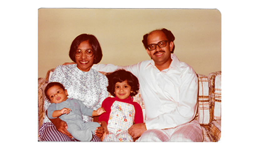 A mother holds a baby and a child sits between the mother and father on a couch.
