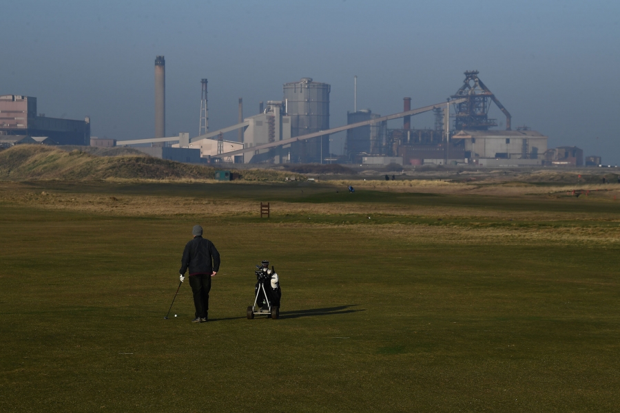 A man plays golf in front of a closed-down steelworks plant in Redcar. A man is show walking with a golf bag with an industrial steelworks plant in the background.