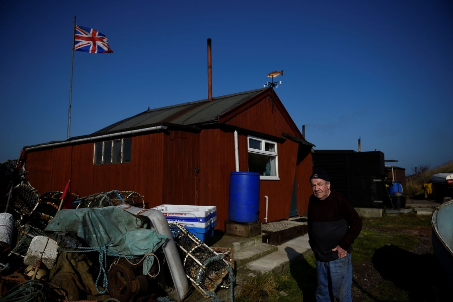 Mohan walks by fishing huts including his own in Redcar. Mohan walks by a red, wooden fishing hut that has a British flag flying behind it.