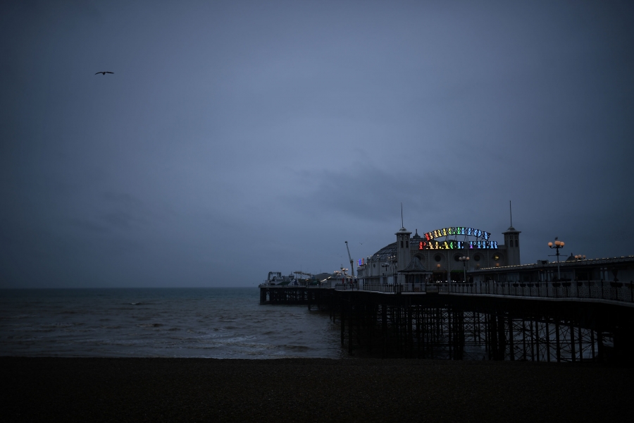 The Brighton Palace Pier sign is illuminated with rainbow colors. A large building is shown at the end of a pier with a sign illuminated with rainbow colors.