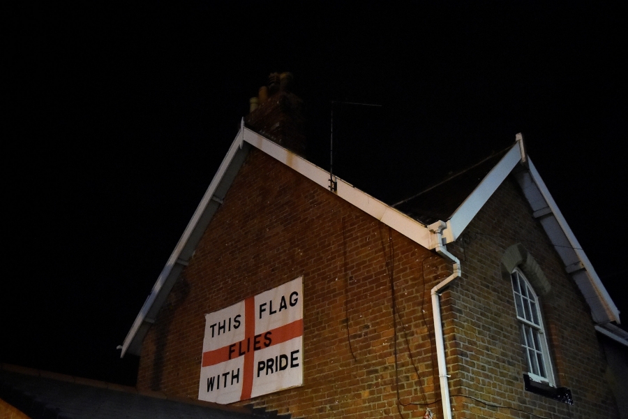 An England flag bearing the words: 'This flag flies with pride' is affixed to the side of a house in Redcar, Britain. An England flag bearing the words: 'This flag flies with pride' is affixed to the side of a red brick house with white trim.