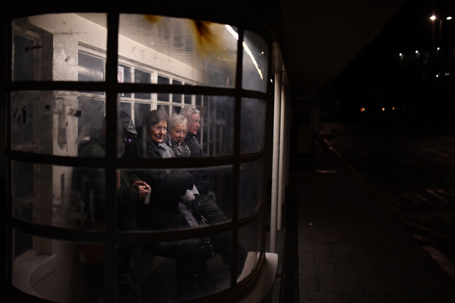 People wait at a bus stop at night in Brighton, Britain. Several older people are shown leaning against the wall of a glass bus stop.