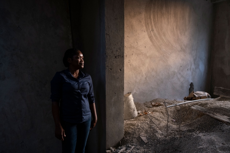 Louise stands inside her home outside Port-au-Prince, Haiti, that she and her family are building with the help of remittances from her cousin, a TPS holder living in Boston. A woman is shown standing among the construction materials of her home with cement walls.