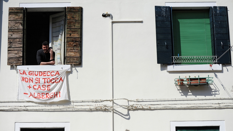 Alessandro Dus, 34, talks on the phone in his illegally occupied apartment in the Casette neighborhood in Venice. Alessandro Dus is show leaning out of a second story open window with a phone in his left hand.