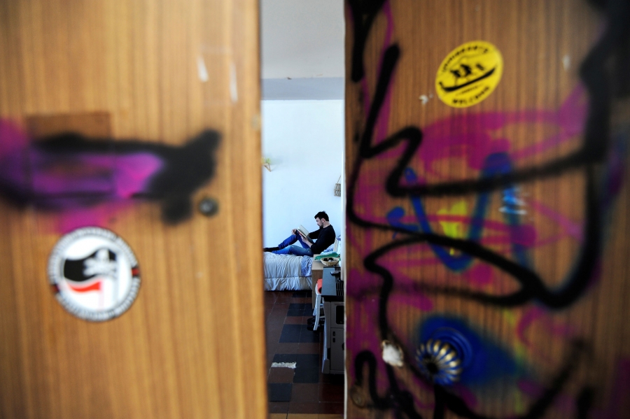 Alessandro Dus reads a book in his illegally occupied apartment. Alessandro Dus is shown through graffiti-painted doors, sitting on a bed reading a book.