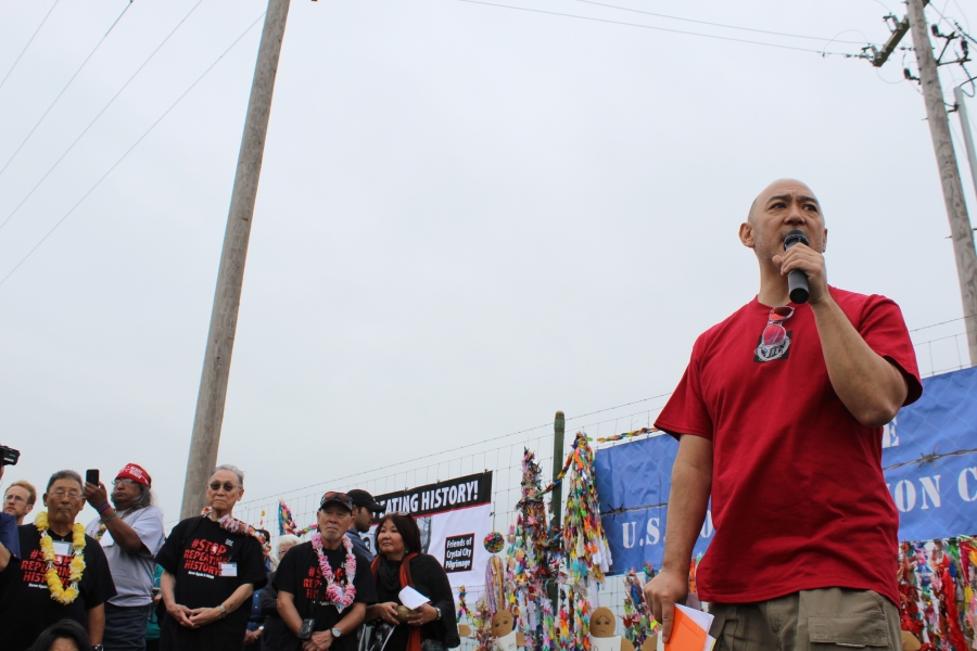 A man stands with a microphone addressing a crowd, with a fence in the background.