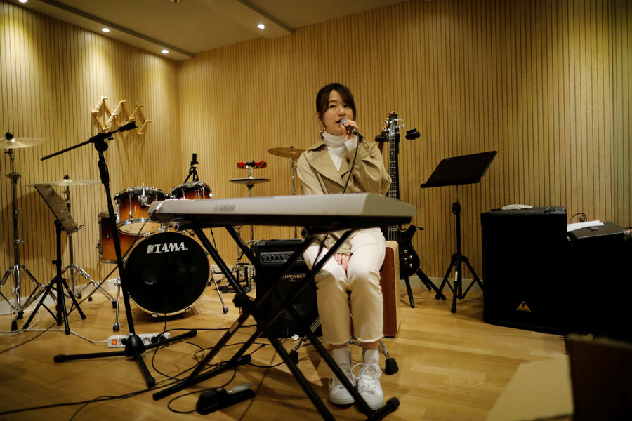 Takeuchi sings during a training session. A woman is shown sitting behind a keyboard and with a microphone in her hand.