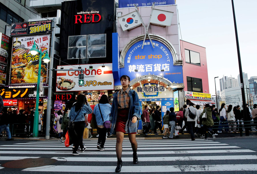 Niitsu walks through Shin-Okubo district, which is known as Tokyo's Korea Town, in Tokyo. A woman is shown walking across a large street with billboard-clad buildings in the background.