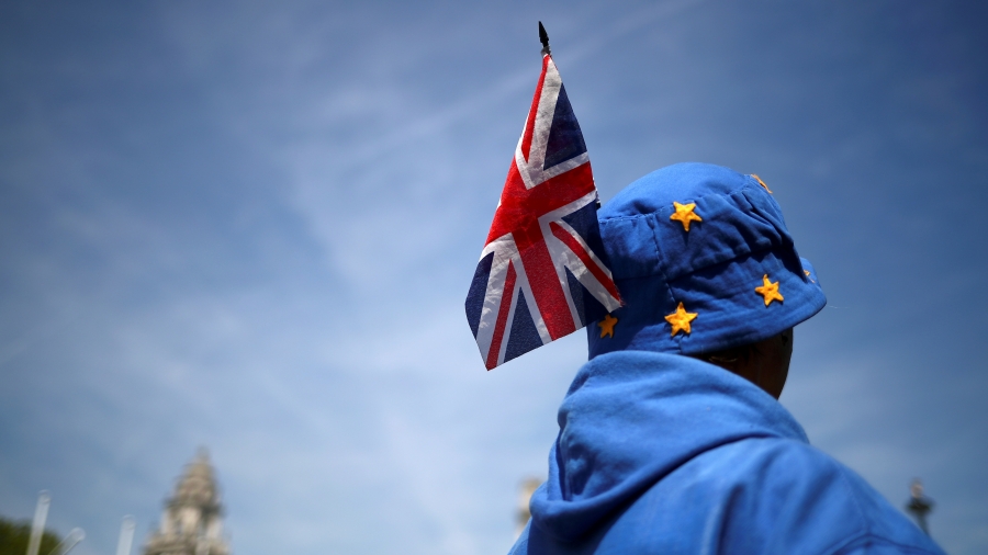 A man in blue with stars on a cap and a UK flag in his had stands with his back facing the camera.