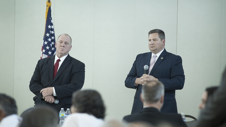 Two men in suits standing in front of room, white wall, with American flag at side