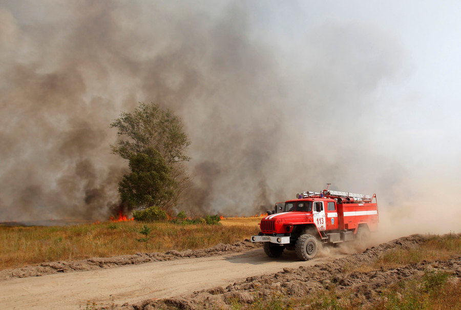 A fire engine drives along a road while smoke from a wildfire cloud the air outside the settlement of Kustarevka in Ryazan region, about 210 miles southeast of Moscow, August 10, 2010. A red fire truck is shown driving across the frame with smoke and fire in the background.