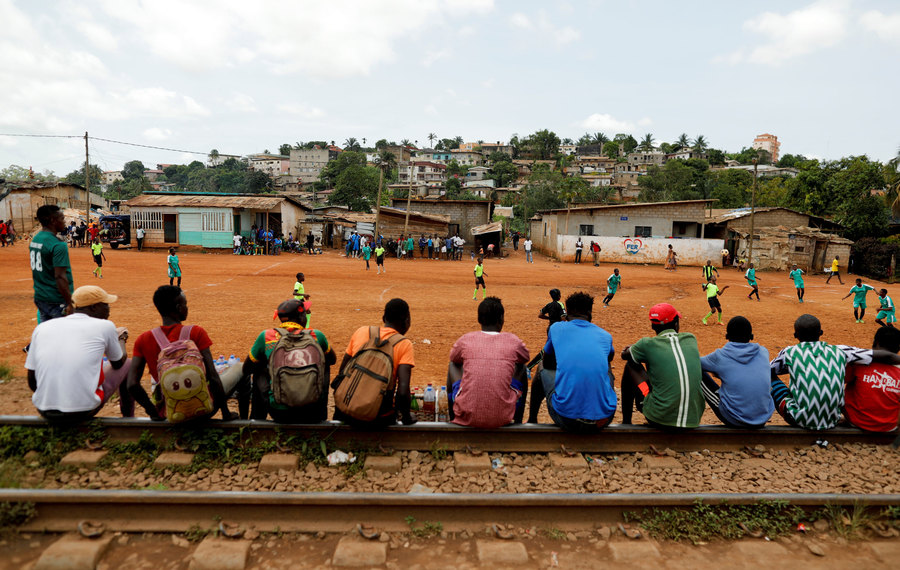 People watch a friendly match between boy's team U15, and girls' team U17. A row of people are shown sitting on railroad track with a soccer match taking place in the distance.