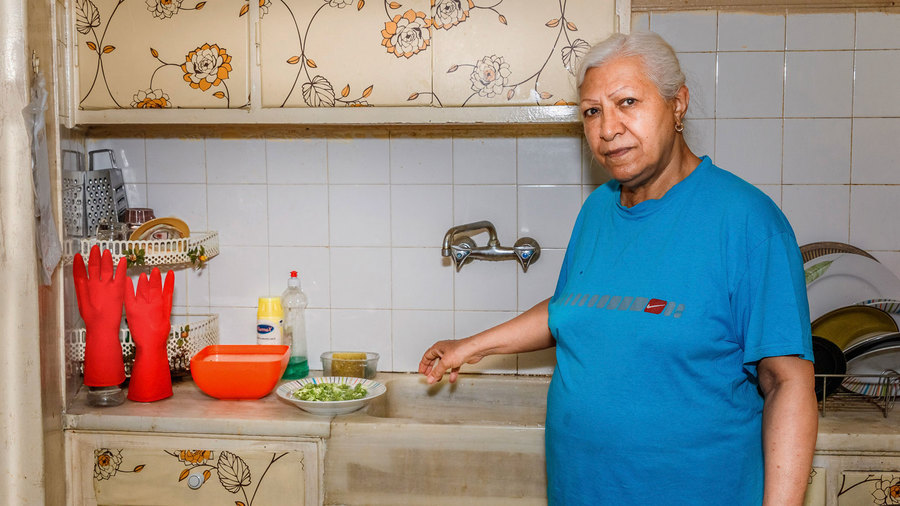 Fatemeh Khani Hakani, 70, helps her daughter by cooking the afternoon meal. Fatemeh Khani Hakani is shown standing next to a sink and flower-lined cupboards.
