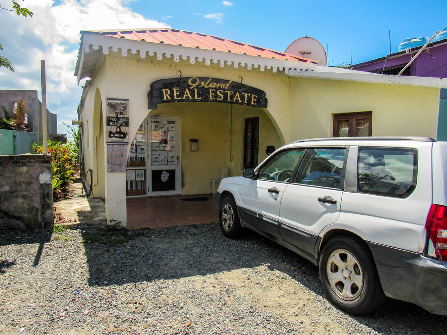 Island Real Estate, one of several real estate agencies on Vieques. A Suburu car is parked outside of a yellow stucco building with a wooden sign that says "Island Real Estate on it."