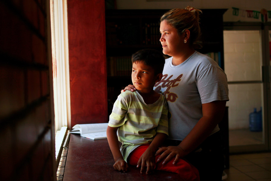 Denia Carranza and her son, are shown at the Casa del Migrante migrant shelter in Ciudad Juarez, Mexico. A woman is shown with her son sitting in front of her while they are both looking out of a window.