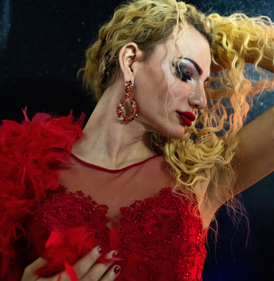 A contestant in the beauty pageant poses for a portrait. A trangender woman is shown with long blonde hair and wearing a red sequin dress.