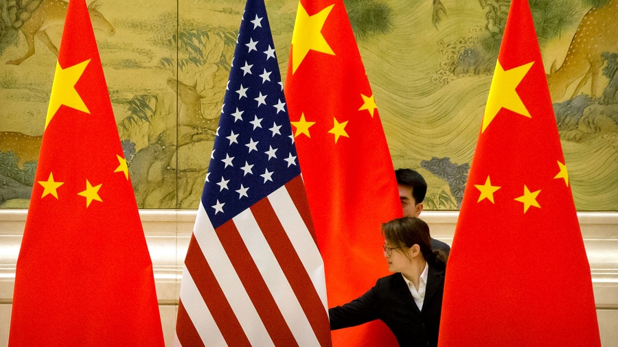 US and Chinese flags are adjusted before the opening session of trade negotiations between US and Chinese trade representatives in Beijing, 2019. One US and three Chinese flags are shown being straightened by a woman standing in the middle of them.