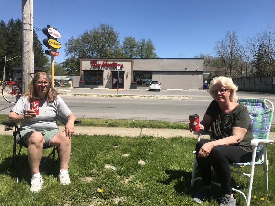Lorrie Chute (left) and Beth Jackson (right) enjoy a coffee in Crystal Beach, Ontario, Canada, practicing the recommended social distancing measures to curb the spread of the coronavirus. Two women sit in lawn chairs and drink a coffee near a coffee shop.