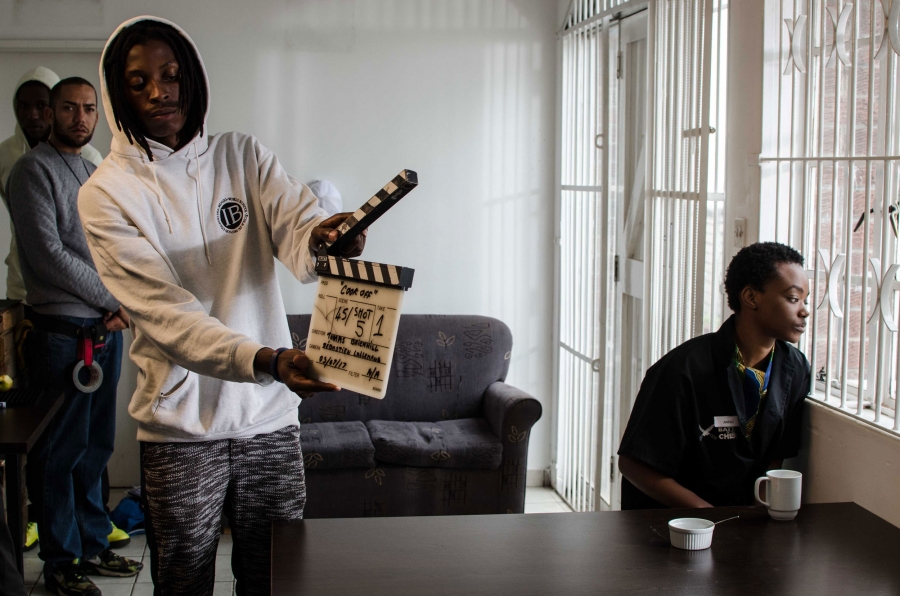 Tendie Chitima, who plays Anesu, on the set of "Cook Off." A man holds a clapperboard as a woman sits at a table looking out a window on a movie set.