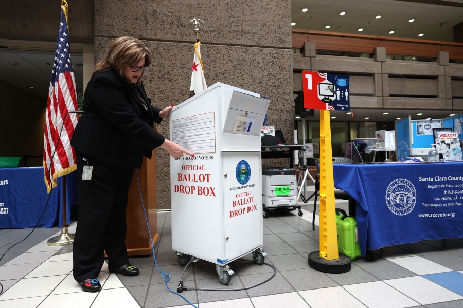 Santa Clara County Registrar of Voters’ Shannon Bushey shows an official county ballot collection box on Oct. 13, 2020 in San Jose, California. A woman stands next to a ballot box, pointing at specific details.