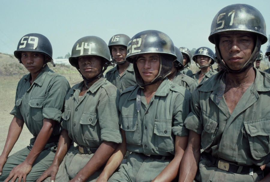 Salvadoran Army recruits await their turn during a parachute training exercise overseen by the U.S. in 1982, during the Salvadoran Civil War. Four women dressed in Army green and helmets sit side by side and make eye contact with the camera.