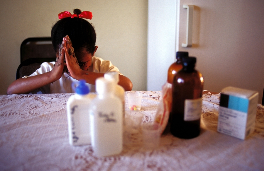 A 9-year-old girl in Johannesburg, South Africa, prays before taking her twice-daily HIV medications in 2002. A young girl folds her hands in prayer in front of her. Out of focus, in front of her, is a group of medication bottles.
